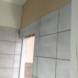 My House recovery - Ściana łazienki z szarymi, kwadratowymi płytkami ceramicznymi, częściowo ułożonymi nad ościeżnicą drzwi. Nad płytkami widoczny fragment ściany przygotowany pod gładzie, sufit biały. W tle widoczne...