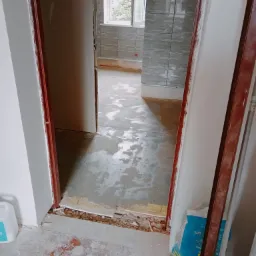 Widok przez futrynę na remontowaną łazienkę z położonymi szarymi płytkami na ścianach, mokrą wylewką na podłodze i workiem zaprawy budowlanej przy ścianie.