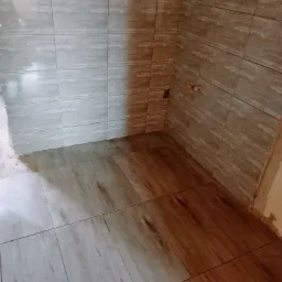 Układanie płytek podłogowych imitujących drewno oraz szarych płytek ściennych w trakcie prac wykończeniowych w rogu łazienki, widoczne wystające kable i rury.
