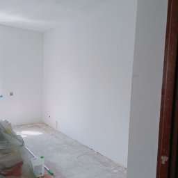 My House recovery - Wnętrze pokoju po świeżym malowaniu na biało, z widocznymi śladami prac na podłodze zabezpieczonej folią, wiadro z zaprawą i butelka płynu do czyszczenia pod ścianą, surowe wykończenie ościeżnicy.