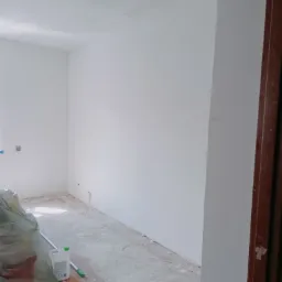 Wnętrze pokoju po świeżym malowaniu na biało, z widocznymi śladami prac na podłodze zabezpieczonej folią, wiadro z zaprawą i butelka płynu do czyszczenia pod ścianą, surowe wykończenie ościeżnicy.