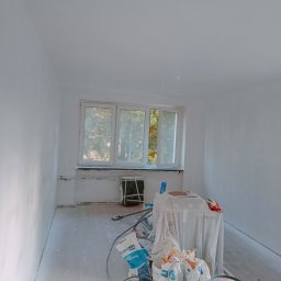 My House recovery - Pokój w trakcie prac wykończeniowych: świeżo pomalowane na biało ściany, okno z widokiem na drzewa, worki z gładzią szpachlową i puszka farby na podłodze przykrytej folią malarską.