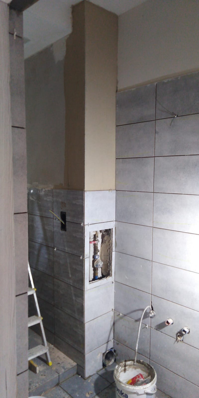 Remont łazienki w trakcie: szare płytki, częściowo położone, odsłonięte rury wodne, drabina aluminiowa w rogu, kubeł z resztkami zaprawy na podłodze.