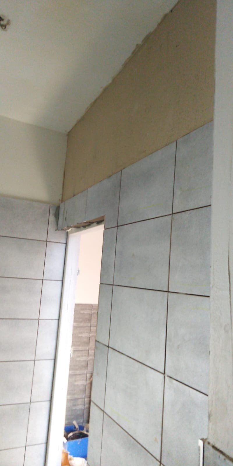 Ściana łazienki z szarymi, kwadratowymi płytkami ceramicznymi, częściowo ułożonymi nad ościeżnicą drzwi. Nad płytkami widoczny fragment ściany przygotowany pod gładzie, sufit biały. W tle widoczne...