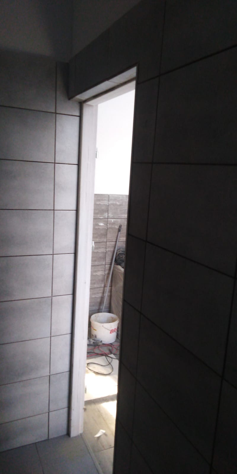 Fragment łazienki w trakcie wykańczania z widoczną ścianą wyłożoną szarymi płytkami, białą futryną drzwi, wiaderkiem po farbie i fragmentem mopa w tle.