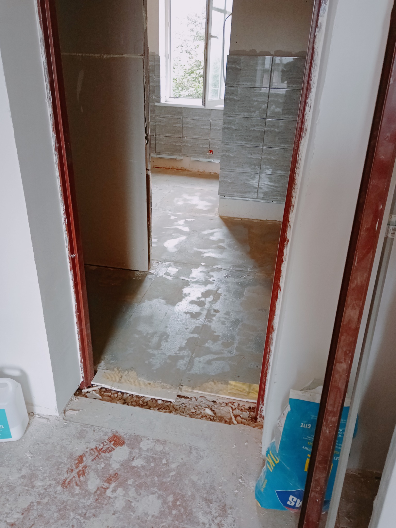 Widok przez futrynę na remontowaną łazienkę z położonymi szarymi płytkami na ścianach, mokrą wylewką na podłodze i workiem zaprawy budowlanej przy ścianie.