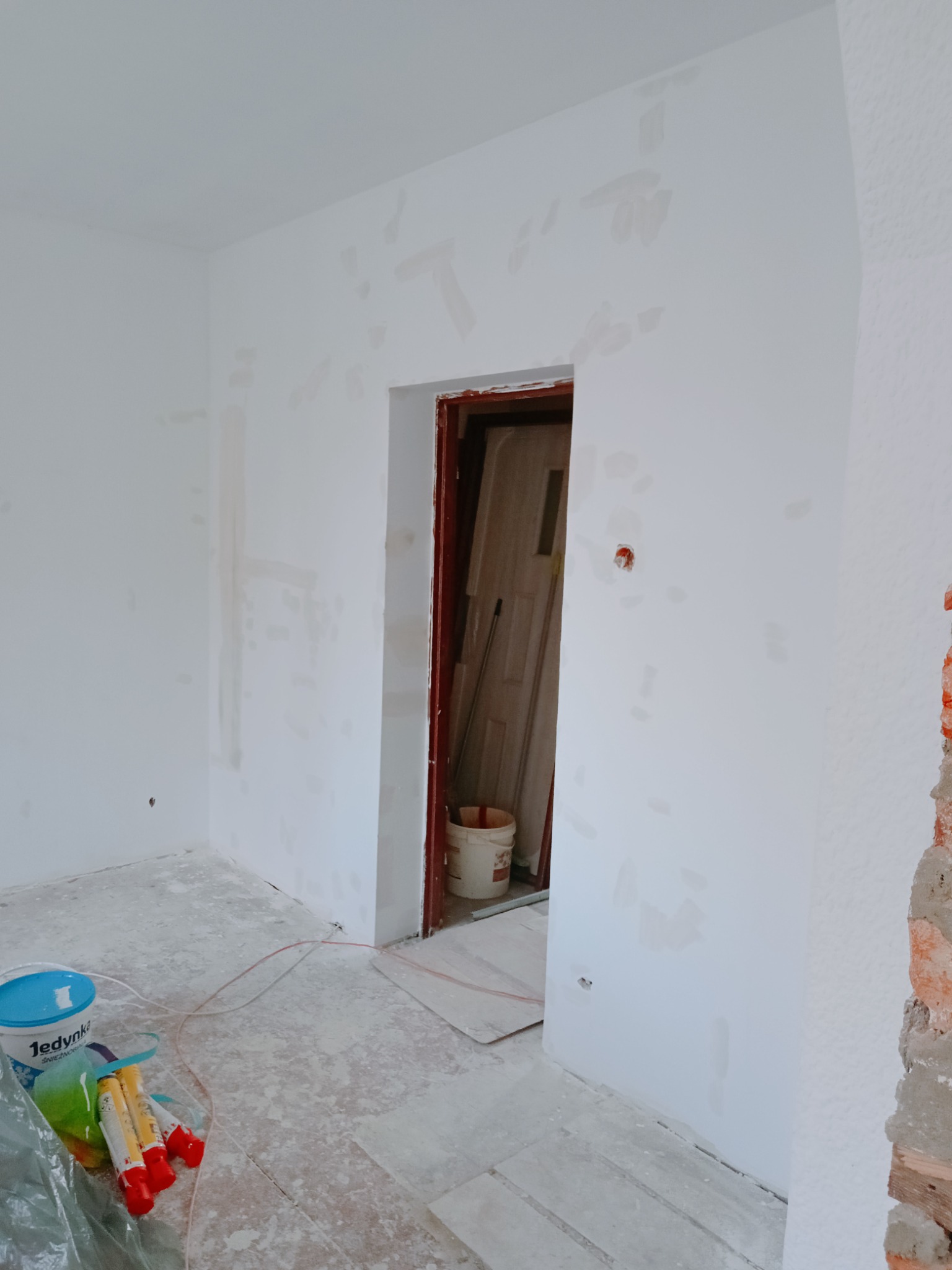 Wnętrze w trakcie przygotowań do malowania w Koszalinie: ściany z widocznymi łatami gładzi, podłoga zabezpieczona folią, wiadro farby Jedynka i narzędzia malarskie w rogu pomieszczenia.