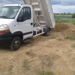 Biała wywrotka z podniesioną skrzynią ładunkową, wysypująca ziemię na pryzmę ziemi, na tle pochmurnego nieba i zieleni.