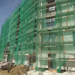 Budynek wielorodzinny w trakcie prac elewacyjnych, osłonięty zieloną siatką na rusztowaniach, widoczne worki z zaprawą i styropian na ziemi.