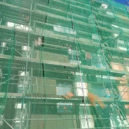 Budynek w trakcie prac elewacyjnych, osłonięty zieloną siatką na rusztowaniach Nova Scaff, widoczne fragmenty elewacji i niebieskie niebo.