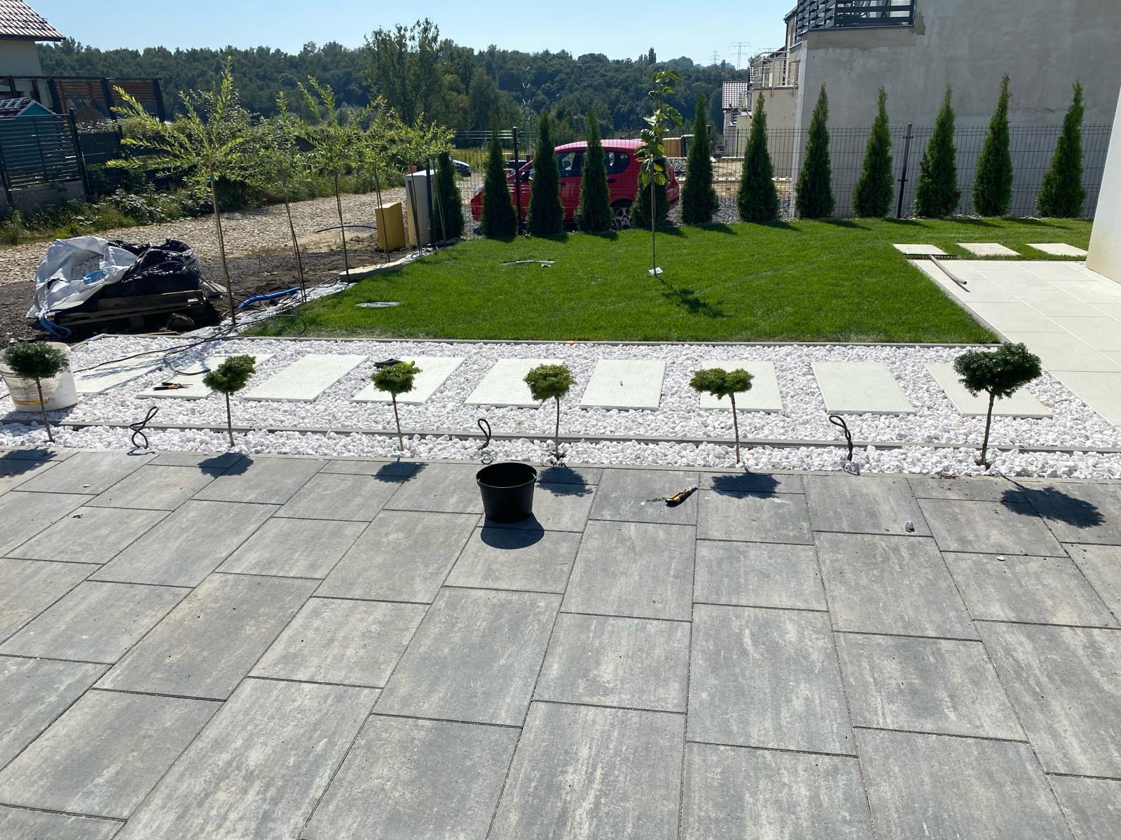 Nowoczesny ogród z szarymi płytami chodnikowymi, białymi kamieniami, ozdobnymi drzewkami i trawnikiem. W tle drzewa i dom.