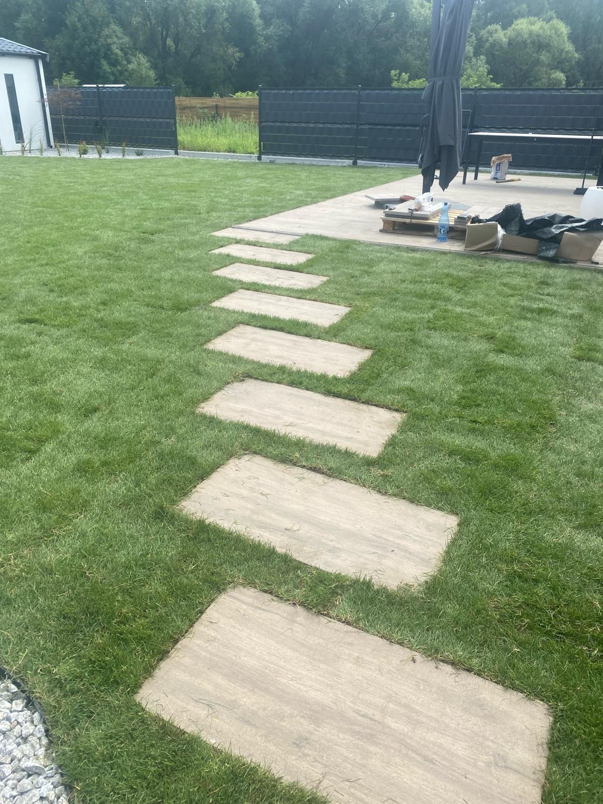 Ścieżka z płyt w trawniku, prowadząca do tarasu z parasolem. Widoczne narzędzia i materiały budowlane. Nowoczesny ogród z czarnym ogrodzeniem.