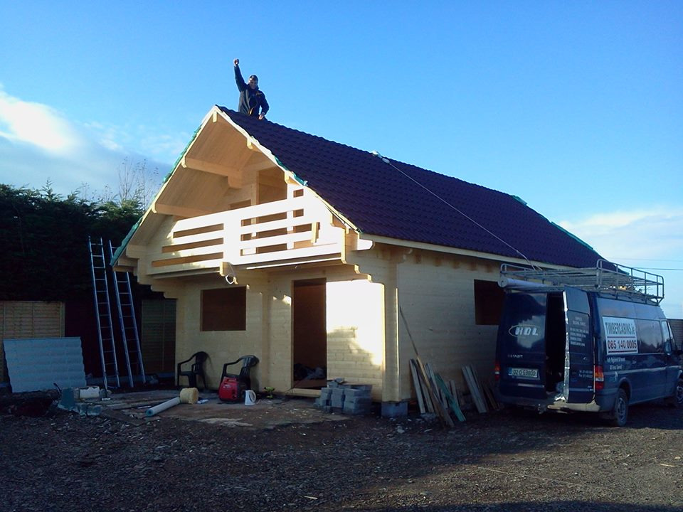 Wznoszenie konstrukcji domu z bali z balkonem; na dachu robotnik z podniesioną ręką, obok zaparkowany bus firmy budowlanej.