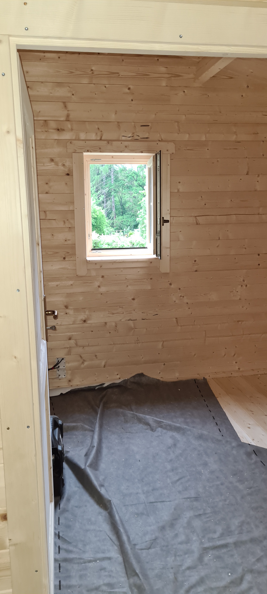 Wnętrze surowego drewnianego domku z bali w trakcie budowy, widoczne okno z widokiem na zieleń, na podłodze rozłożona folia zabezpieczająca.