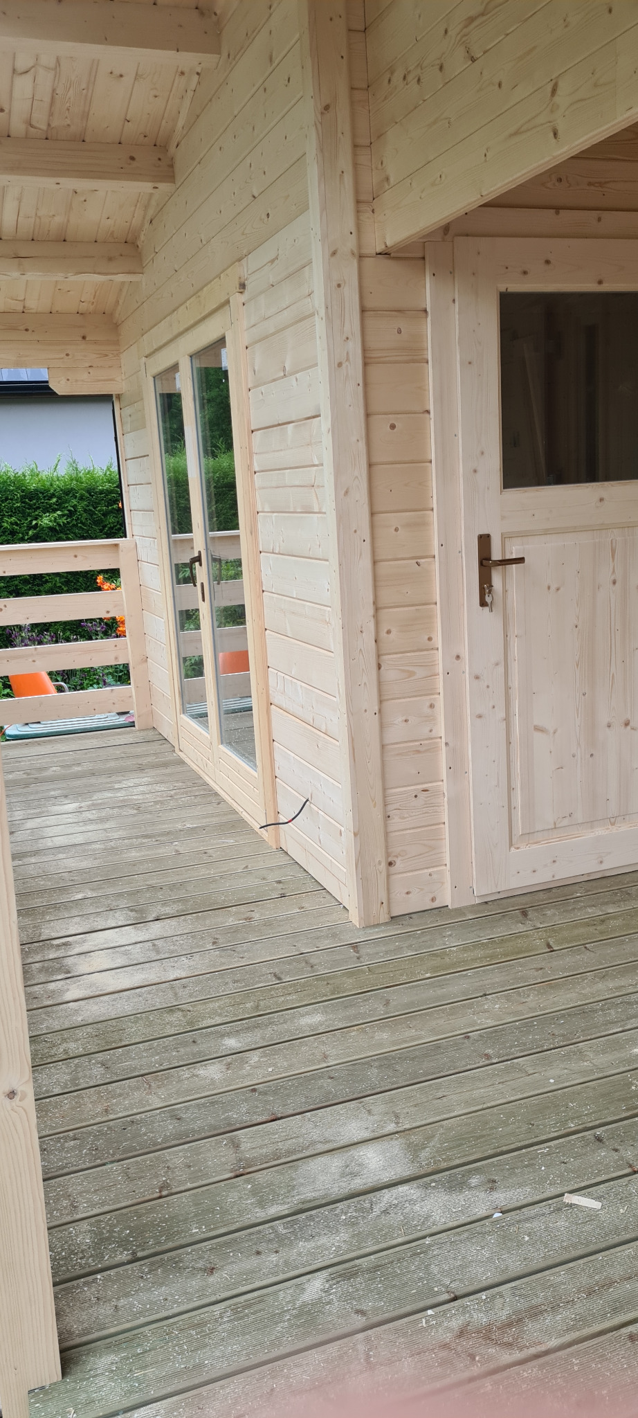 Fragment drewnianej konstrukcji z bali z widocznymi drzwiami z szybami i wejściem na taras z drewnianą podłogą.