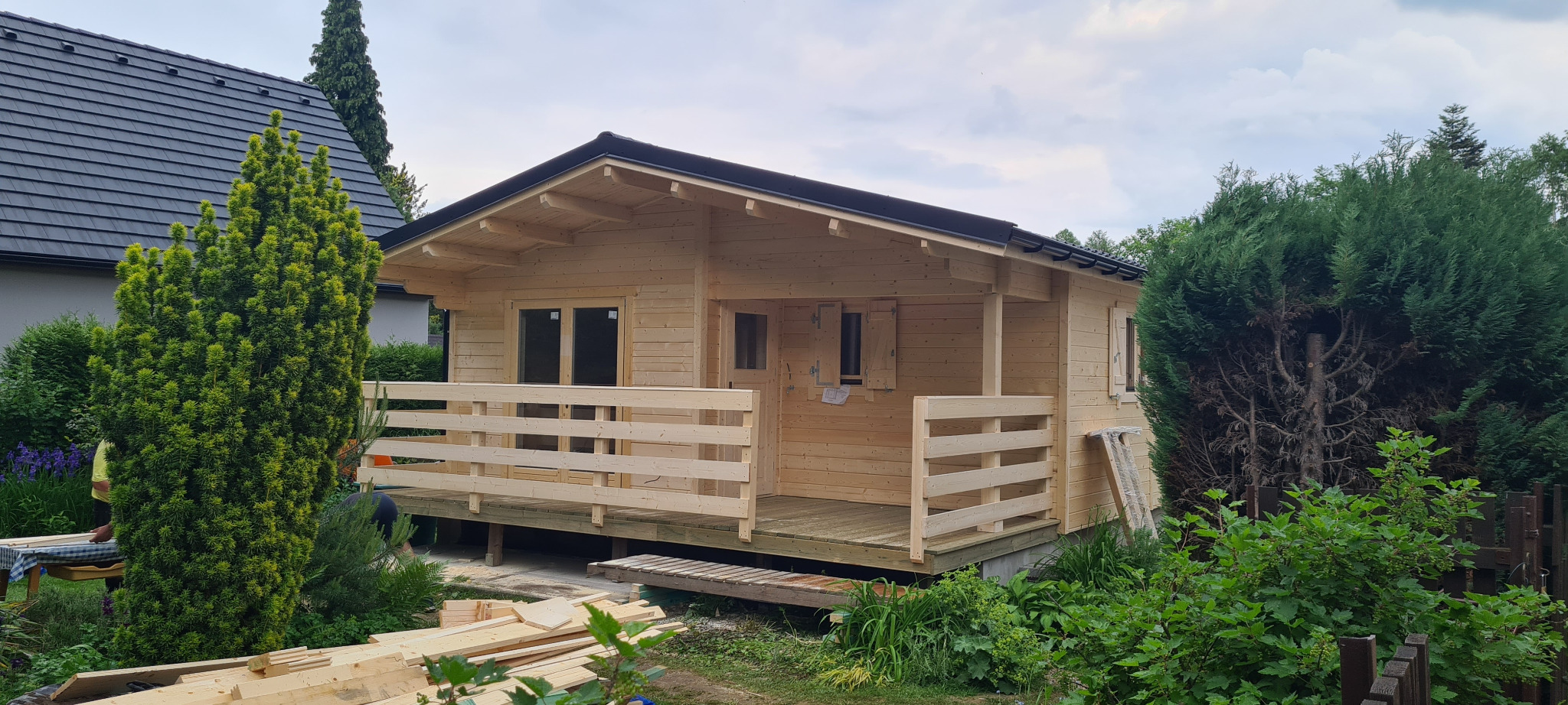 Surowy drewniany domek z bali z tarasem w trakcie budowy, widoczne deski i narzędzia w otoczeniu zieleni.