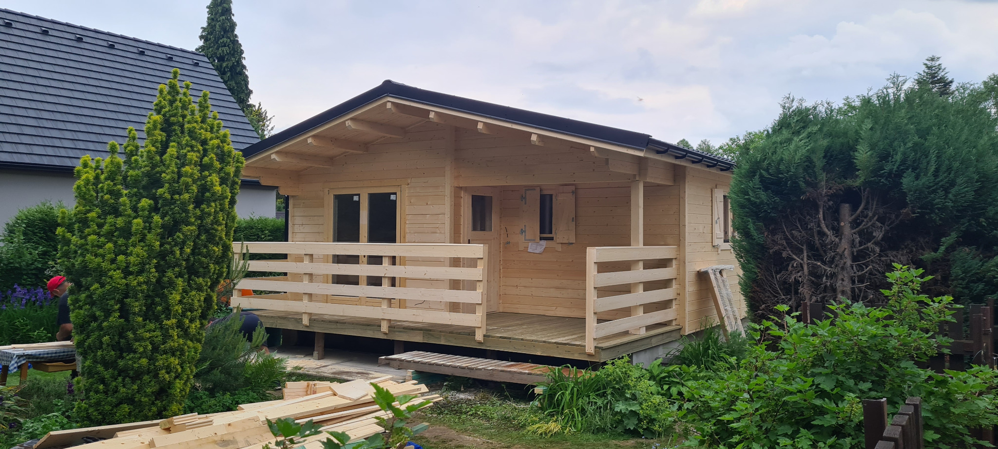 Surowy drewniany domek letniskowy z tarasem, częściowo wykończony, widoczne deski i narzędzia na zewnątrz, w tle dom z ciemną dachówką.