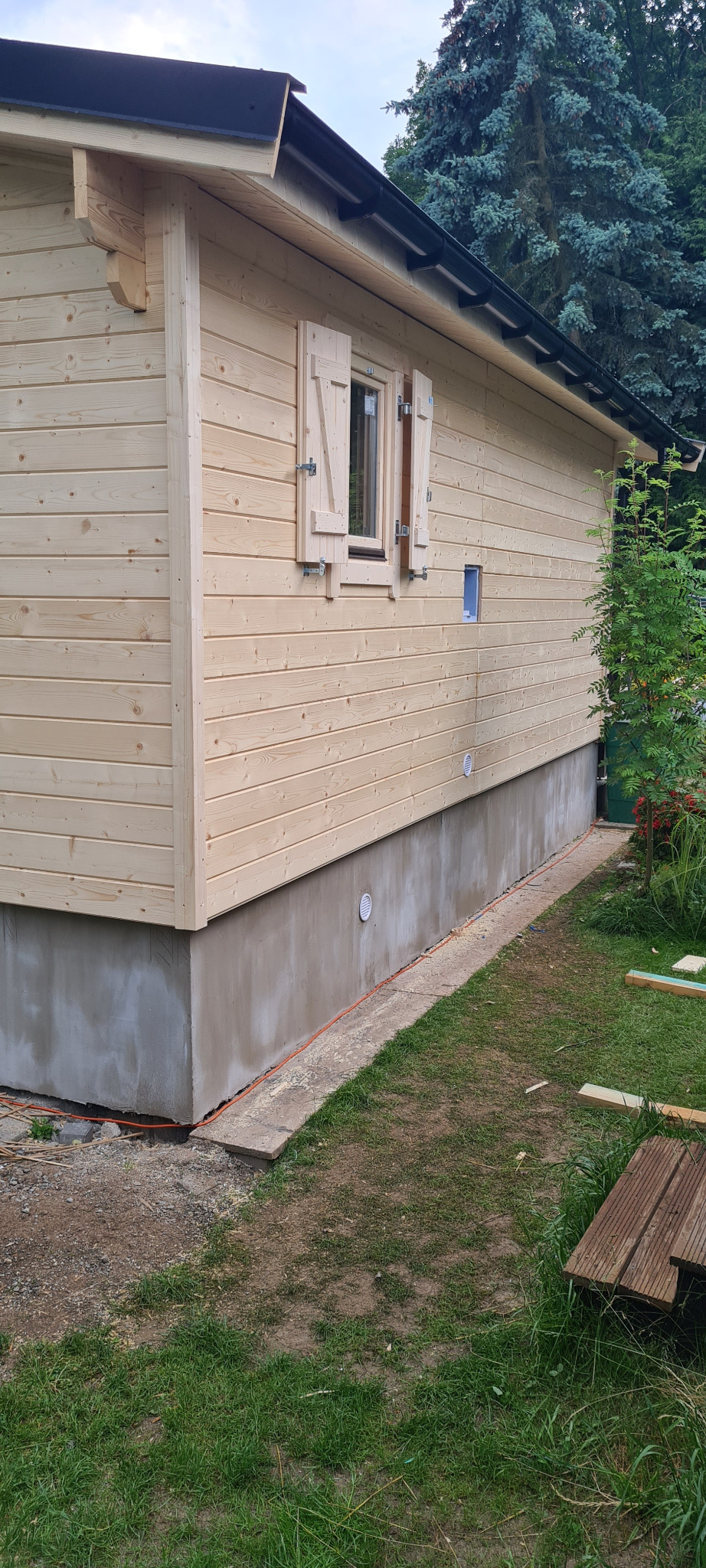 Ściana boczna surowego domu z bali z otwartymi okiennicami, widoczne fundamenty i elementy instalacji elektrycznej na zewnątrz.