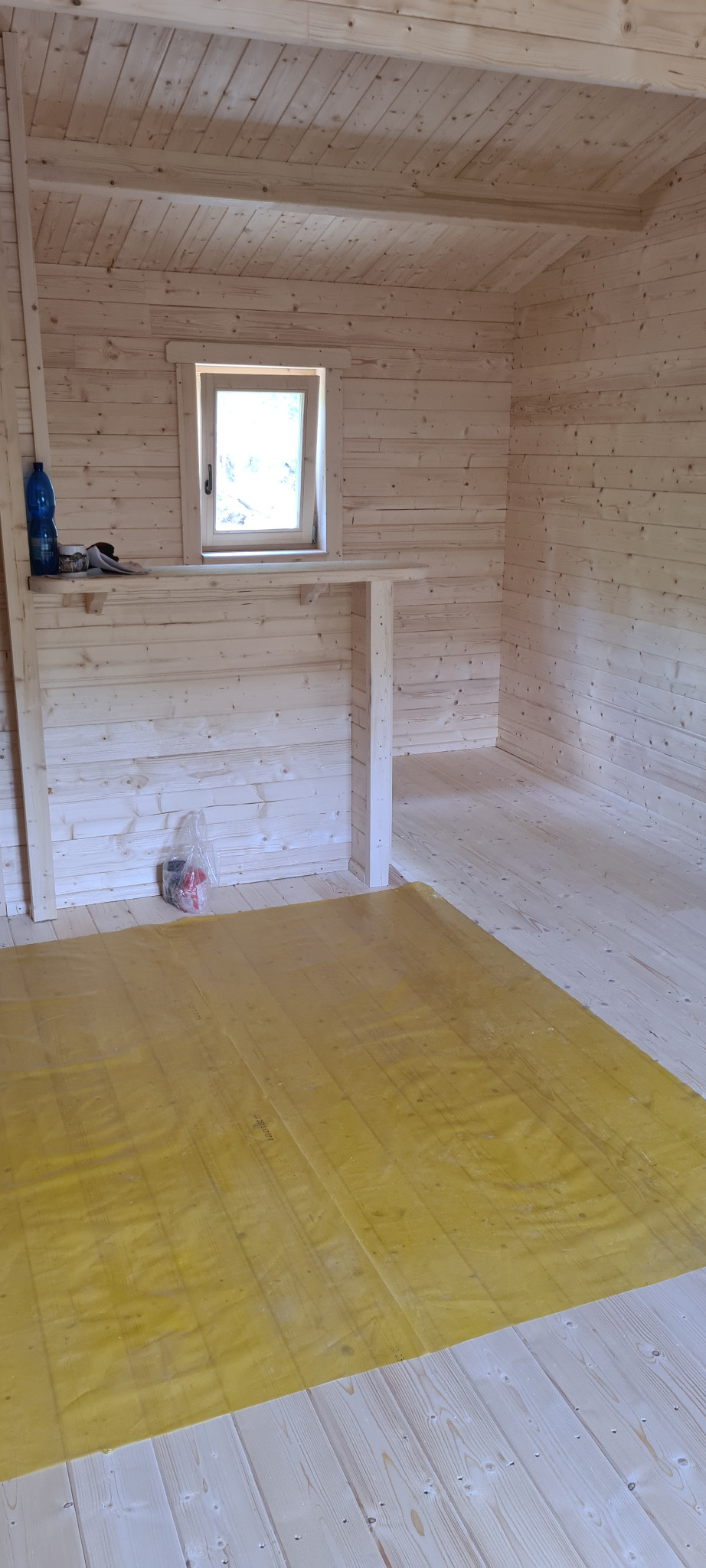 Wnętrze surowego drewnianego domku z bali w trakcie budowy, z żółtą folią ochronną na podłodze, butelką wody i narzędziami na półce pod oknem.