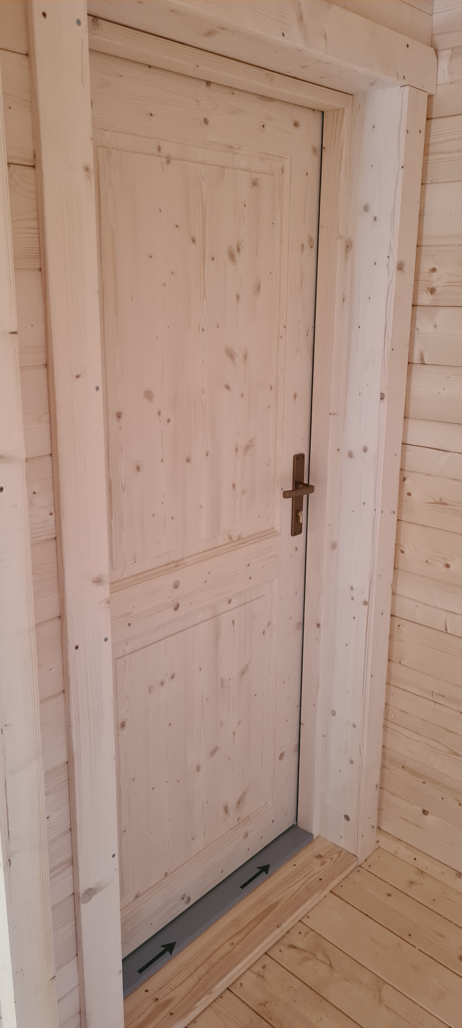 Jasna, drewniana, panelowa wewnętrzna drzwi w domu z bali z widoczną uszczelką pod drzwiami z nadrukowanymi strzałkami.