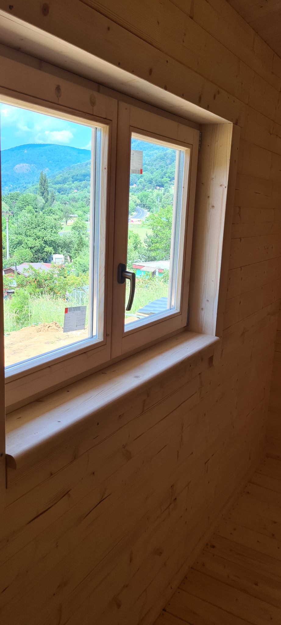 Wnętrze domu z bali: drewniane okno z widokiem na zielony krajobraz z pagórkami.