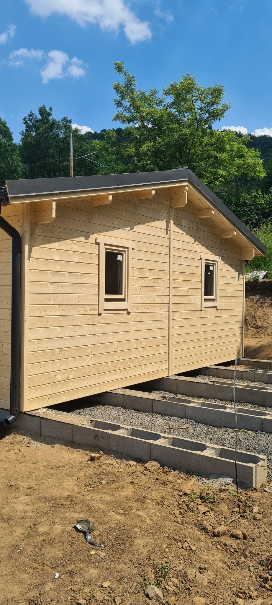Surowy drewniany domek, widok z boku, osadzony na betonowych bloczkach wypełnionych żwirem, w tle zieleń drzew.