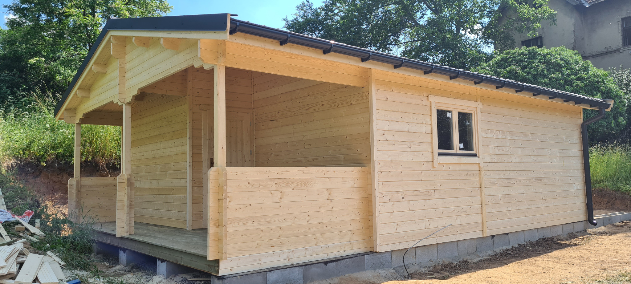 Surowy, jasny drewniany budynek z bali z czarnym dachem i rynnami, widok z zewnątrz, częściowo wykończony, z widocznymi bloczkami fundamentowymi i stertą nieużytych desek obok.