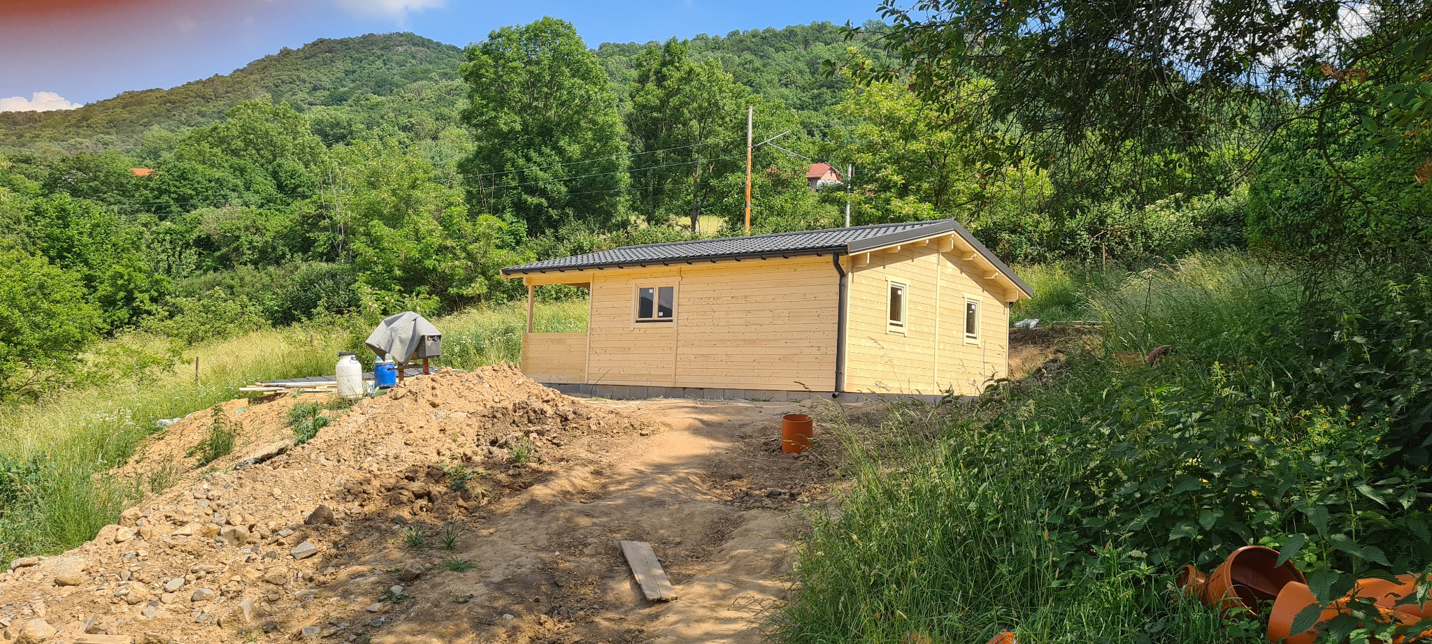 Surowy drewniany domek letniskowy z czarnym dachem na nierównym terenie, otoczony zielenią i przygotowany do dalszych prac wykończeniowych.