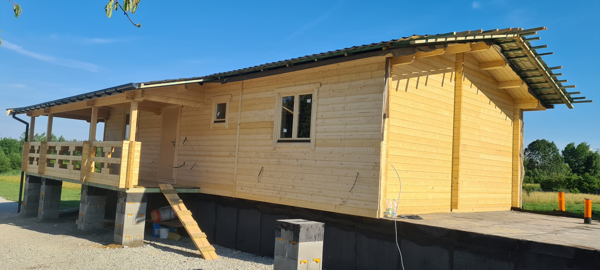 Surowy dom z bali w trakcie budowy, widoczne bloczki fundamentowe, drewniane ściany i częściowo wykończony dach, zarysowany taras i schody wejściowe.