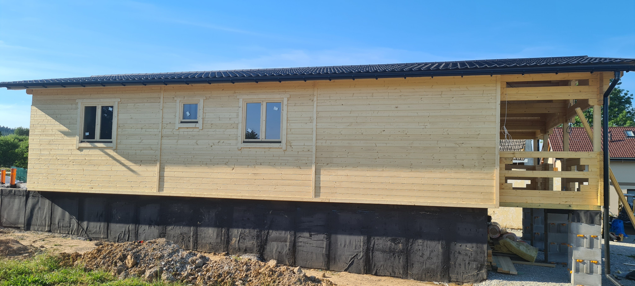 Surowy stan elewacji jasnego, drewnianego domu z bali z czarnym dachem, widoczne okna z folią zabezpieczającą, czarna hydroizolacja fundamentów i częściowo wykonana drewniana konstrukcja werandy.