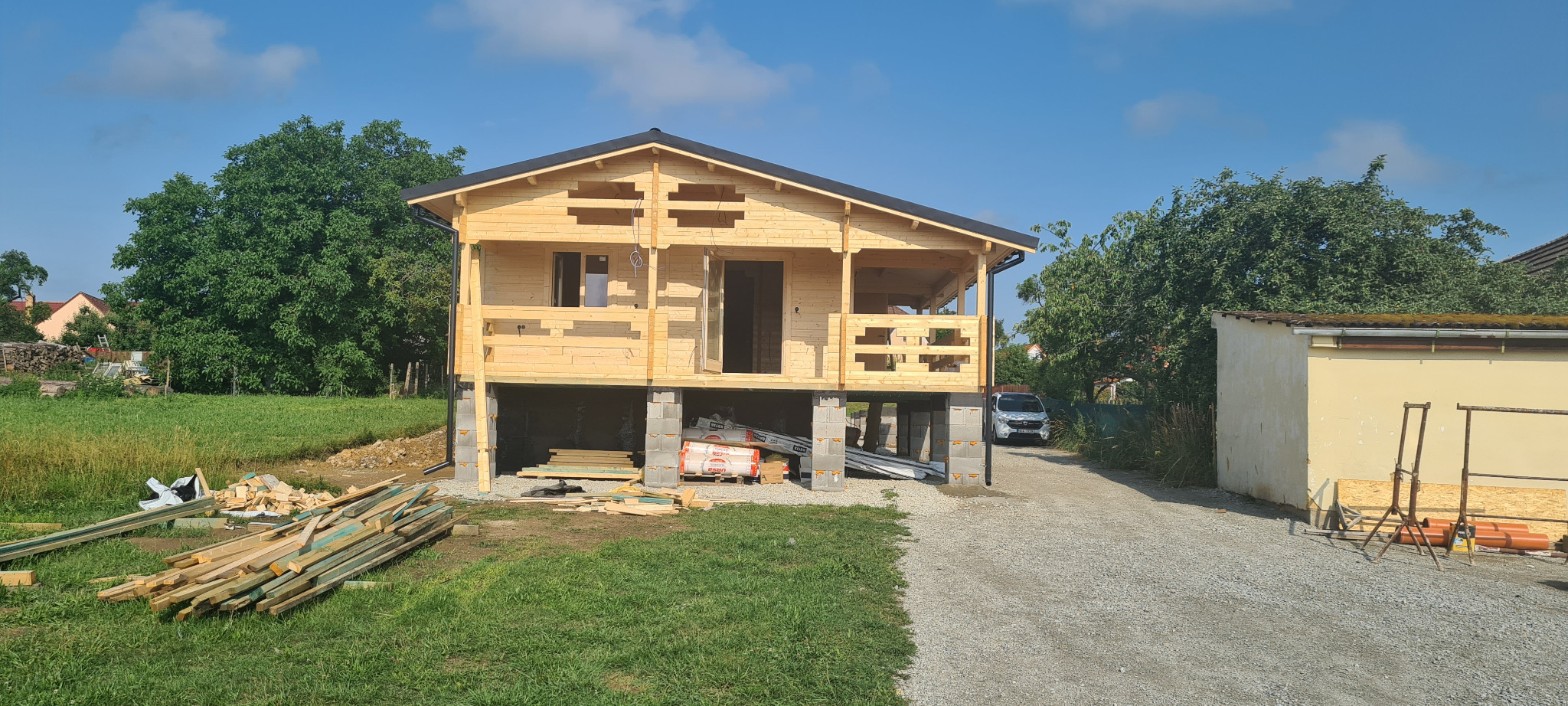 Surowa drewniana konstrukcja domu, posadowiona na betonowych bloczkach, z widocznymi materiałami budowlanymi i zaparkowanym samochodem w tle.