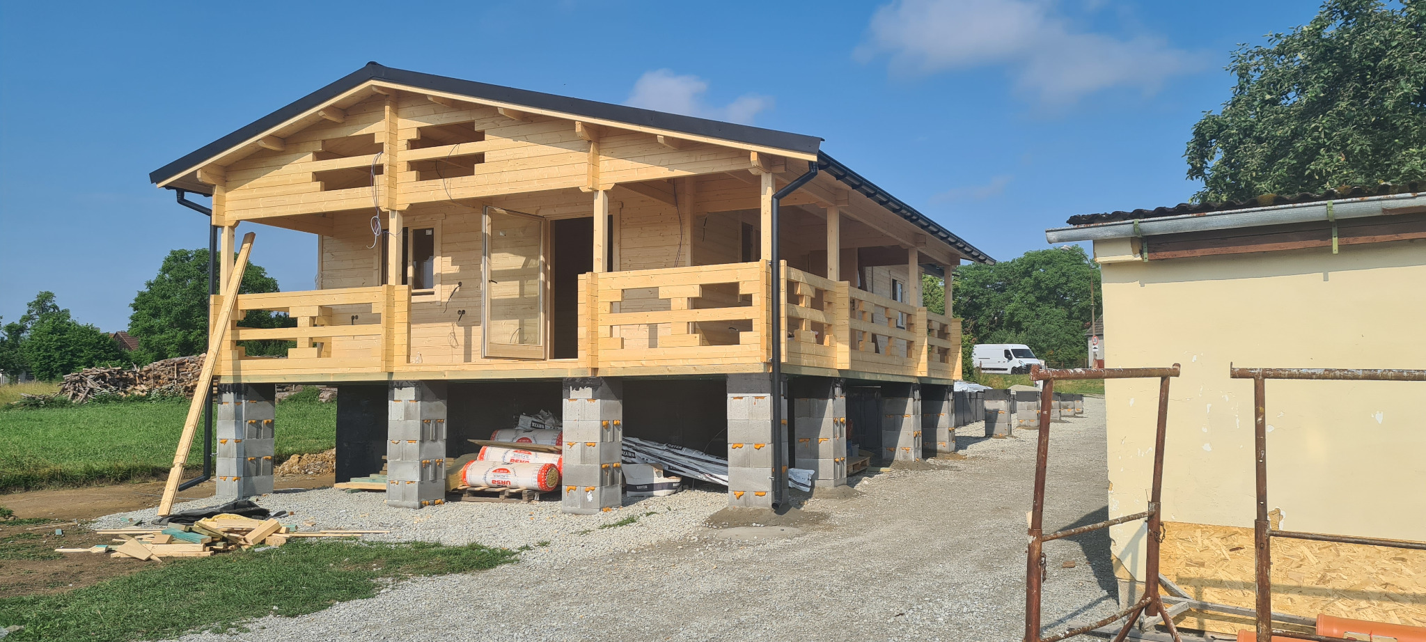 Surowy drewniany dom z bali na betonowych bloczkach, z widoczną izolacją i elementami konstrukcyjnymi, w trakcie budowy, na tle błękitnego nieba i zieleni.
