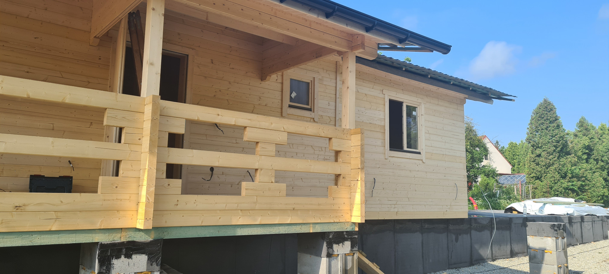 Surowy stan elewacji domu z bali na betonowej podmurówce, widoczne elementy konstrukcyjne werandy i okna, w tle zieleń drzew.