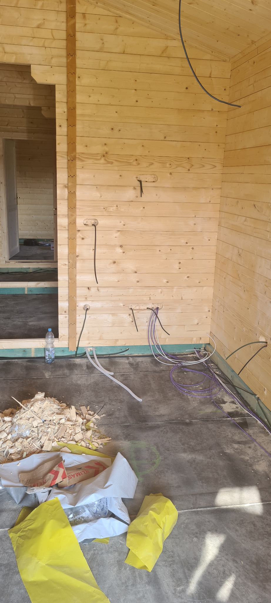 Wnętrze surowego, drewnianego domu w trakcie budowy, widoczne przewody elektryczne w ścianach, woda mineralna i odpady budowlane na podłodze.