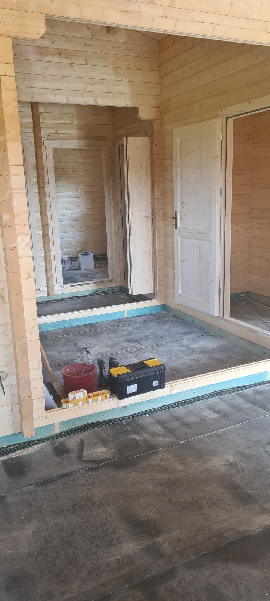 Wnętrze surowego drewnianego budynku w trakcie budowy, widoczne narzędzia, wiadro i skrzynka narzędziowa na podłodze, surowe drewniane ściany i ramy drzwiowe.