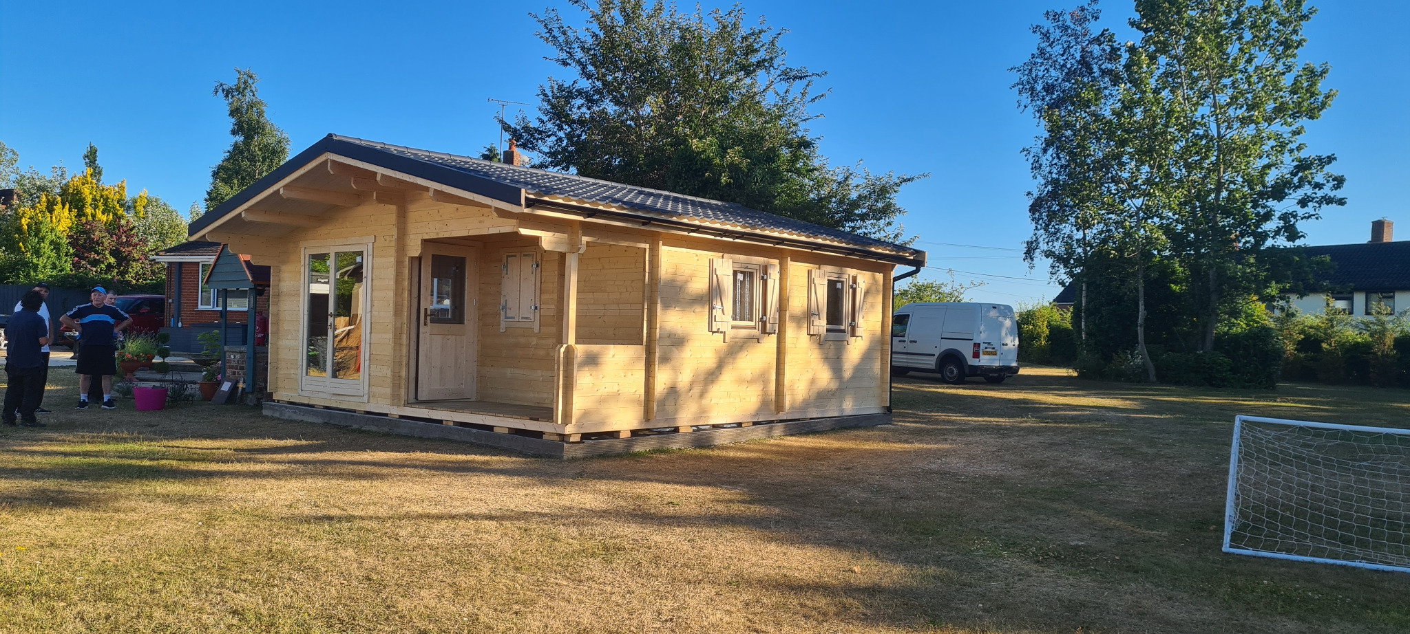 Jasny, parterowy domek letniskowy z bali, z dwuspadowym dachem i małymi okiennicami, ustawiony na trawniku, z grupą osób i białym vanem w tle.