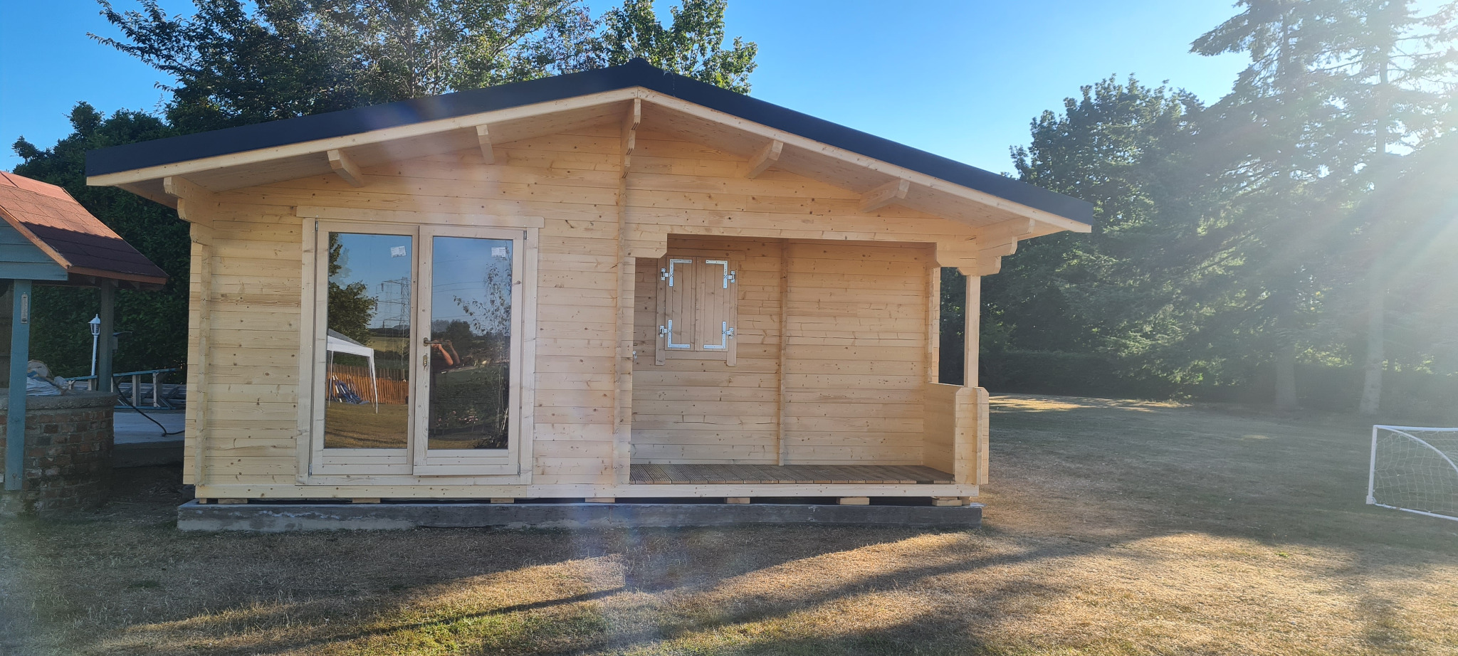 Surowy, drewniany domek letniskowy z bali na fundamencie, z dwuskrzydłowymi drzwiami i małym okienkiem z okiennicą, w tle zieleń drzew i fragment zabudowań.
