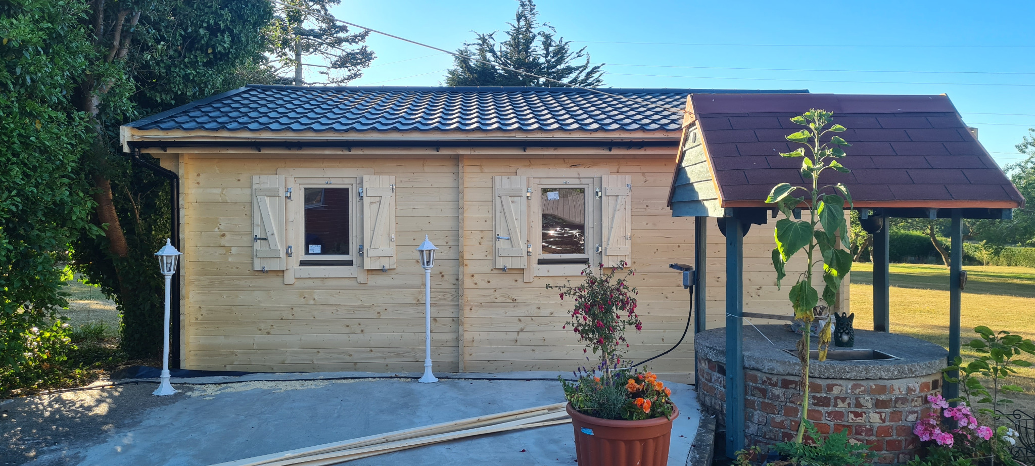 Dwuokienne domki z bali z czarnym dachem i białymi okiennicami, z dekoracyjną studnią i kwiatami w donicy na pierwszym planie