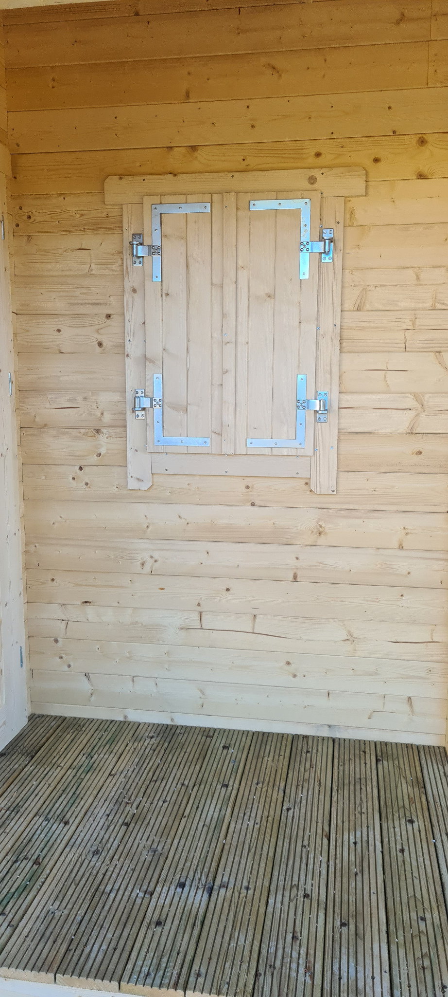 Wnętrze szopy z bali z drewnianą podłogą i zamkniętym okienkiem z metalowymi zawiasami.