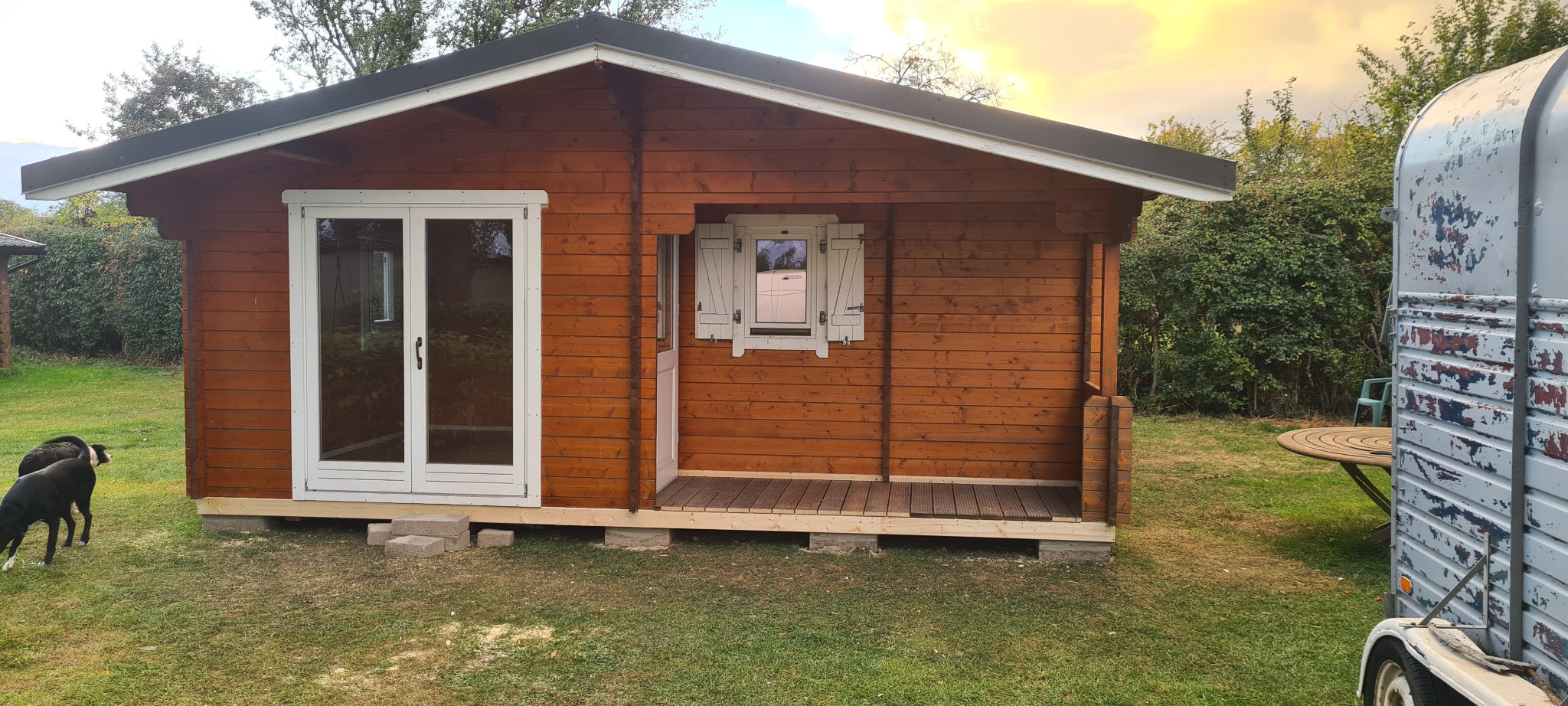Niewielki, brązowy domek z bali z białymi oknami i drzwiami w Nisko, na zielonej trawie, z widocznym psem i przyczepą w tle.
