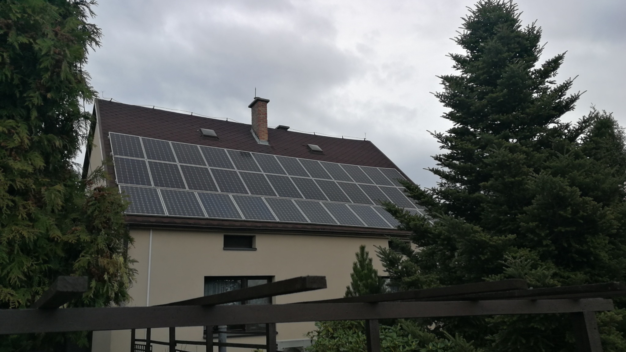 Dom jednorodzinny z panelami słonecznymi na dachu z brązowej dachówki, widoczne drzewa iglaste i pochmurne niebo w tle.