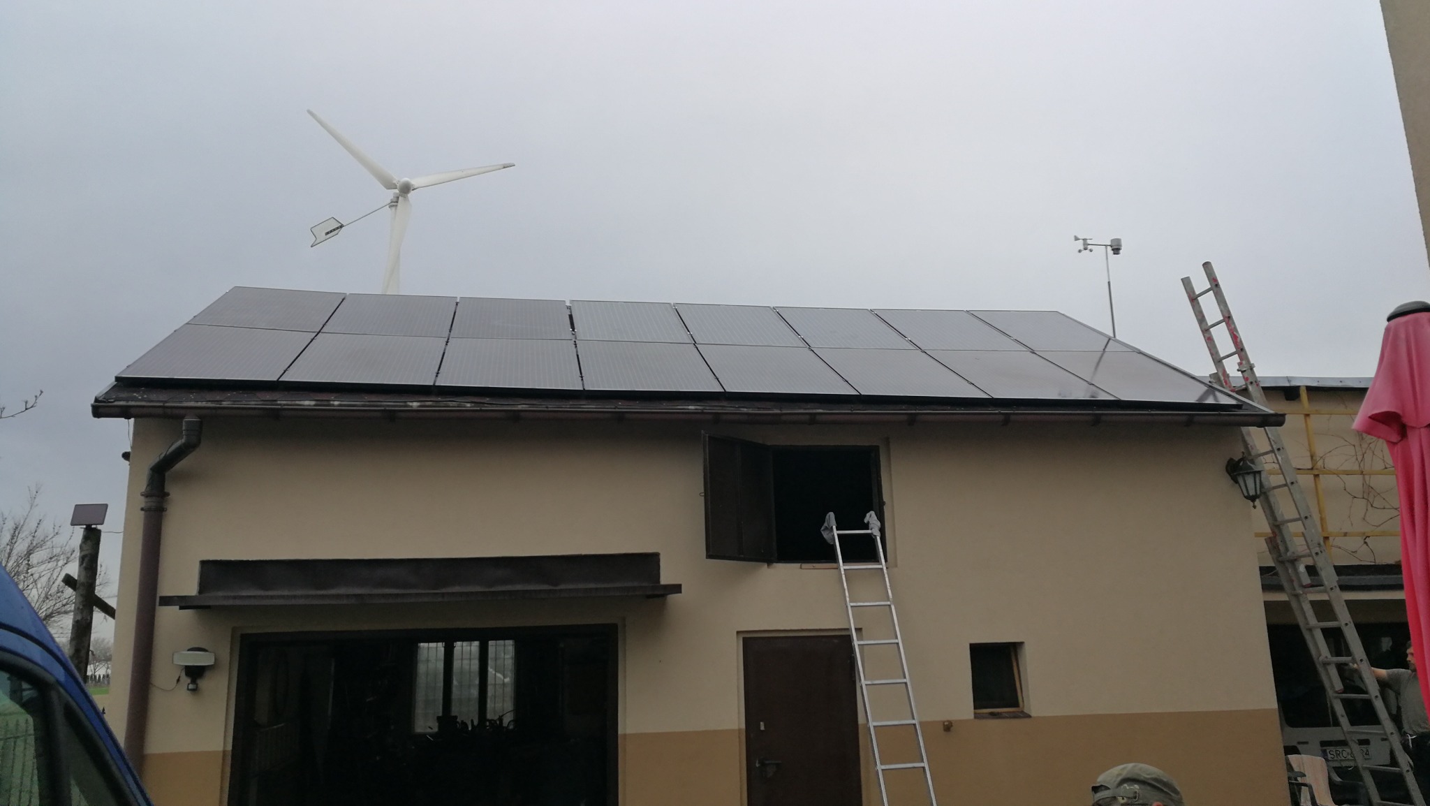 Dom z panelami słonecznymi na dachu i turbiną wiatrową w tle, drabina oparta o ścianę, pochmurne niebo.