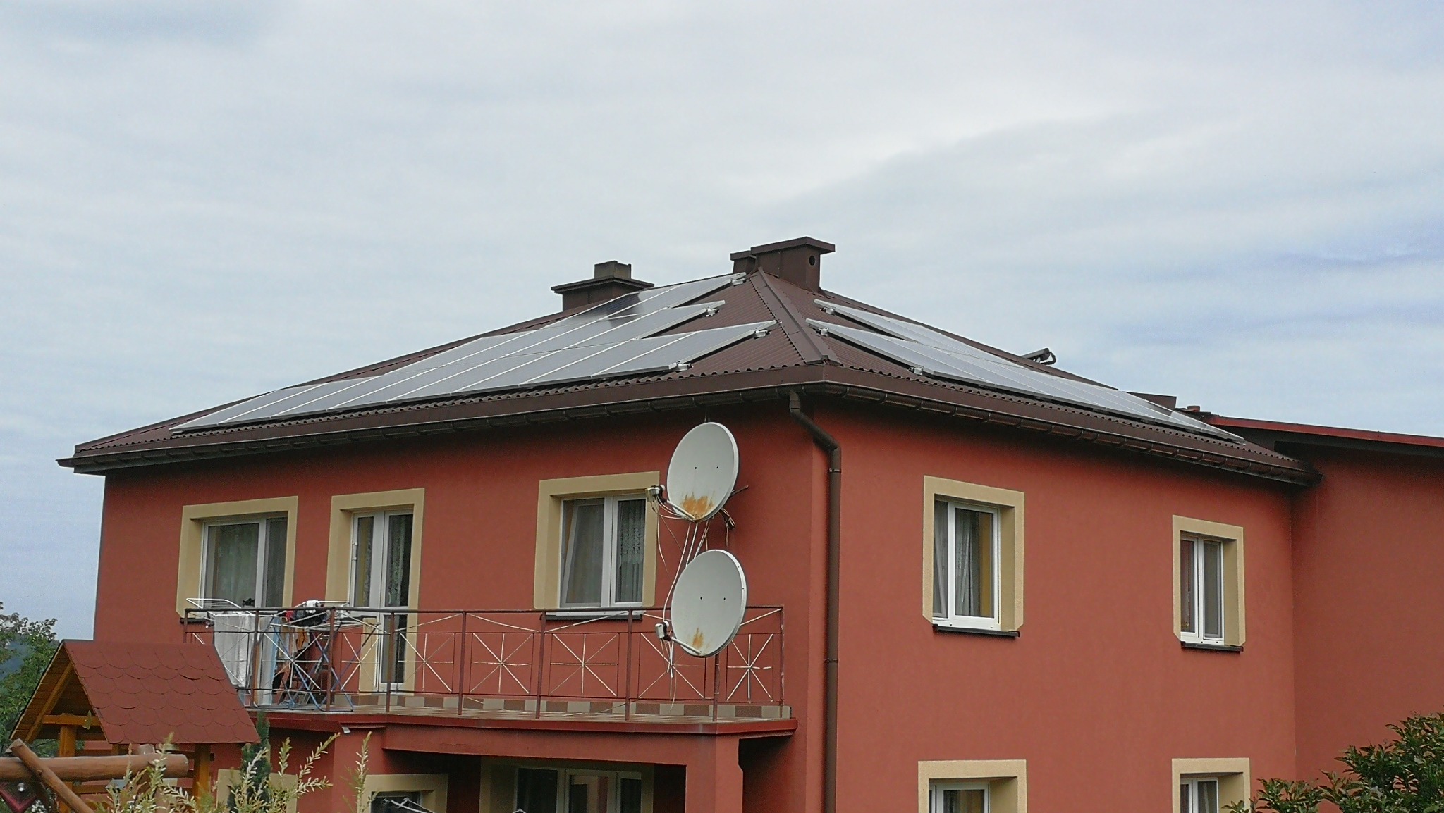 Czerwony budynek z panelami słonecznymi na brązowym dachu, widoczne dwie anteny satelitarne na ścianie i balkon z suszarką na pranie.