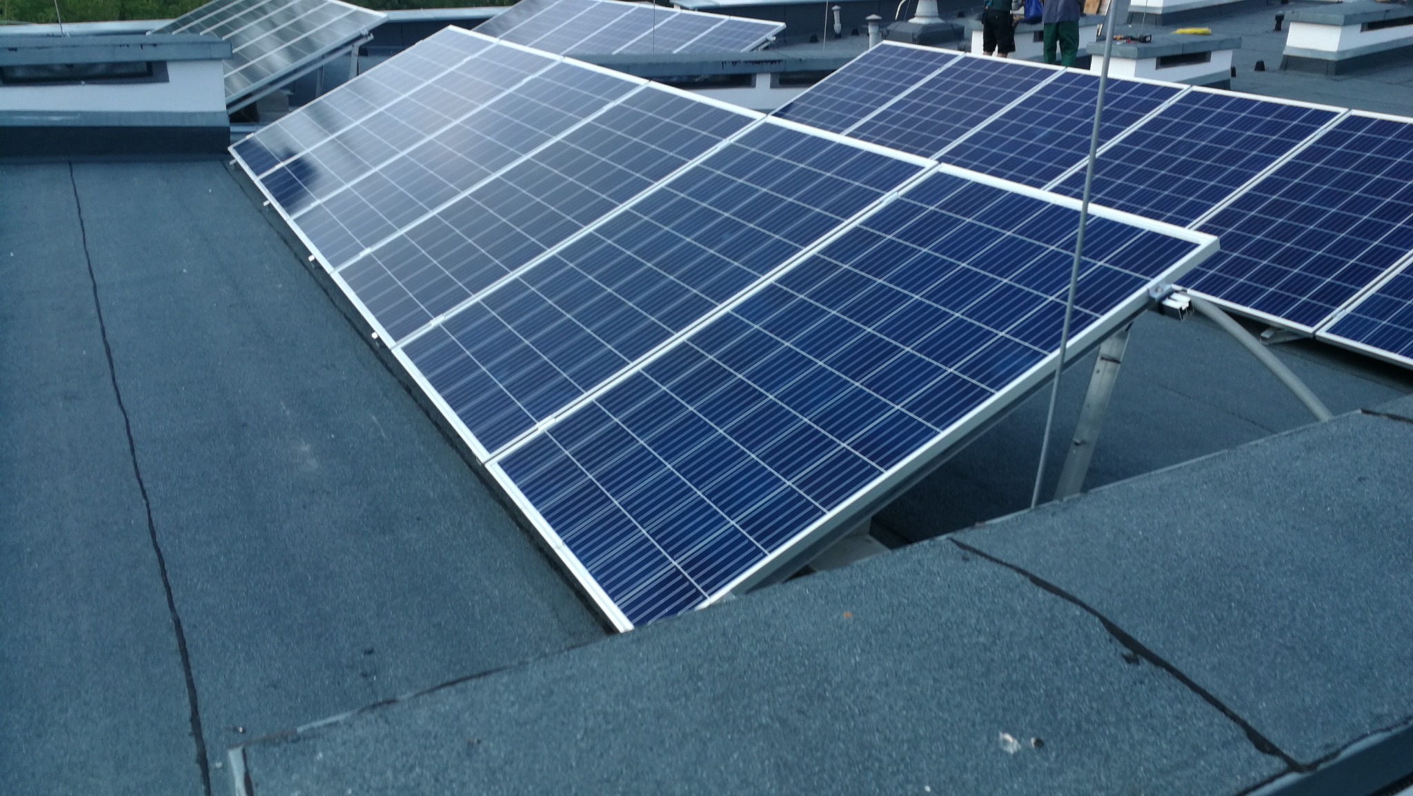 Rząd paneli słonecznych na płaskim dachu pokrytym papą, widoczne metalowe konstrukcje wsporcze i fragmenty sylwetek instalatorów w tle.