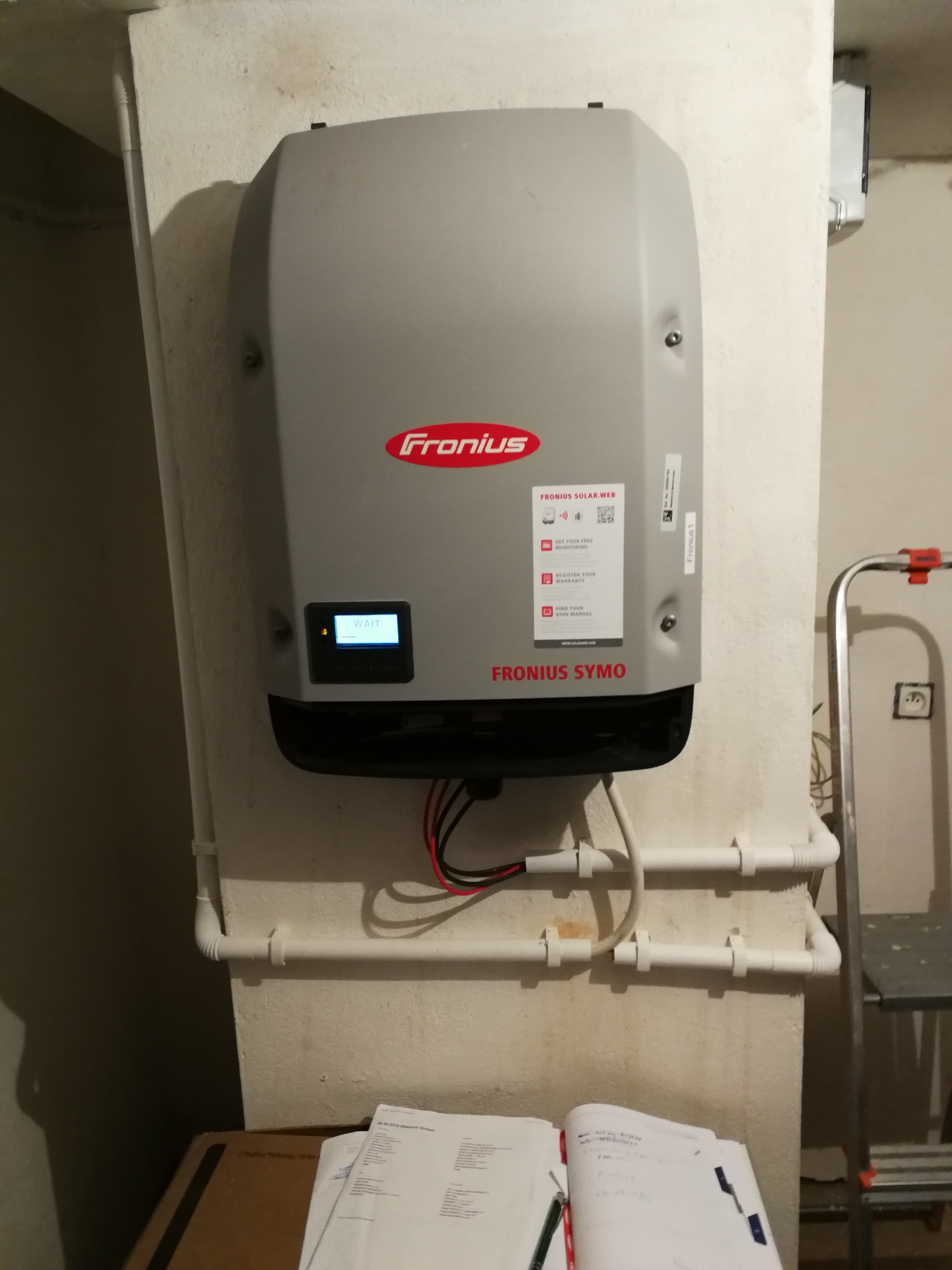 Szary inwerter Fronius Symo zamontowany na ścianie z widocznymi przewodami, rurami instalacyjnymi i notatnikiem leżącym poniżej, ekran inwertera wyświetla napis 'WAIT'.