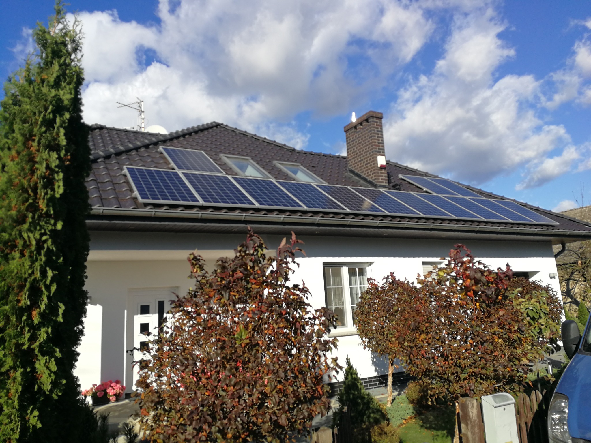 Biały dom z ciemną dachówką, na której zamontowano panele słoneczne; widoczne również okna dachowe, komin z czujnikiem i antena telewizyjna; na pierwszym planie drzewa i krzewy.