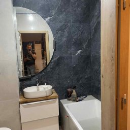 KAM-PRO Kamil Jędrzejewski - Wykończona łazienka z umywalką na szafce, wanną i ciemnymi płytkami imitującymi marmur na ścianie. W lustrze odbija się osoba robiąca zdjęcie.