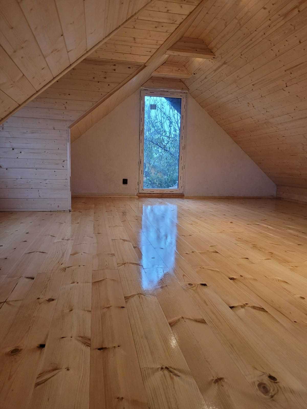 Drewniane wykończenie poddasza: boazeria na ścianach i suficie, lakierowana podłoga z desek, okno z widokiem na drzewa. Naturalne światło podkreśla ciepło drewna.