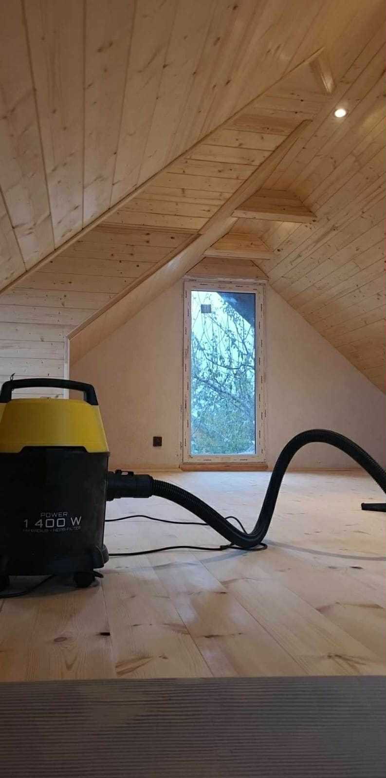 Drewniane poddasze po remoncie z oknem i odkurzaczem przemysłowym. Widoczne panele na podłodze i drewniana boazeria na skośnym suficie. Odkurzacz w kolorze czarno-żółtym.