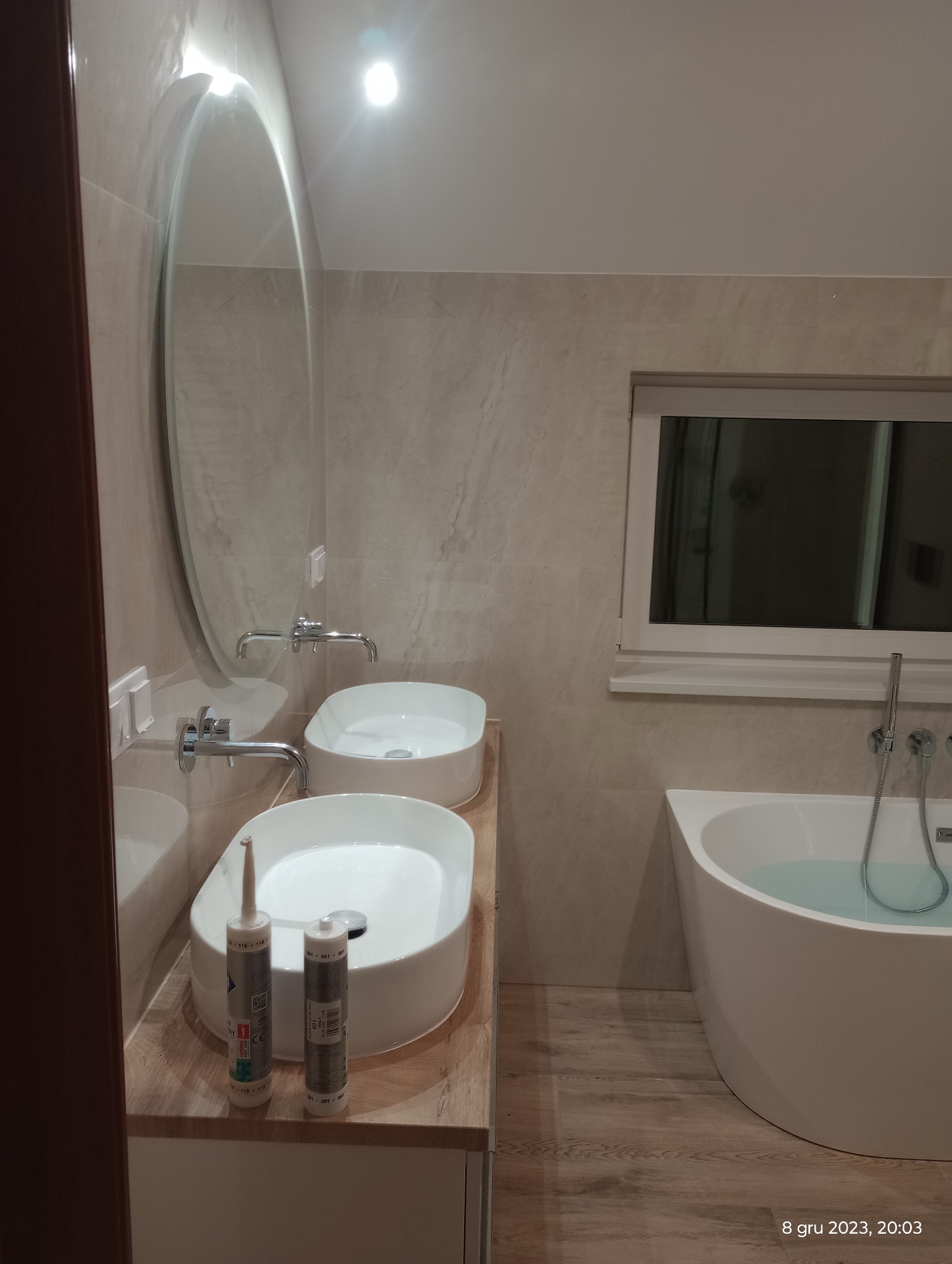 Wykończona łazienka z dwiema umywalkami nablatowymi, owalnym lustrem i wanną. Na blacie tuby silikonu. Płytki imitujące marmur na ścianach i podłodze.